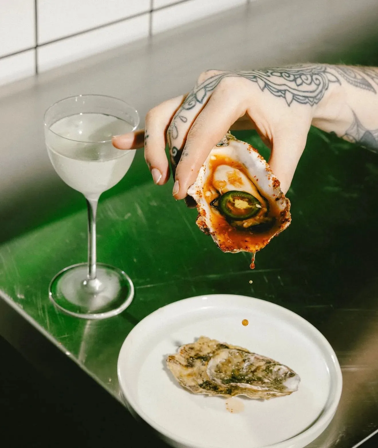 A hand holding a chargrilled oyster with hot sauce, next to a martini glass